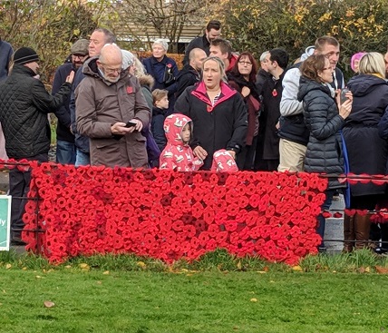 Photo of the General Public in the road & a knitted Poppy Blanket on the Church Fence